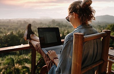 Frau arbeitet remote auf Balkon mit Ausblick Frau sitzt mit Laptop auf Balkon und arbeitet mit Blick über Baumwipfel bei Sonnenuntergang
